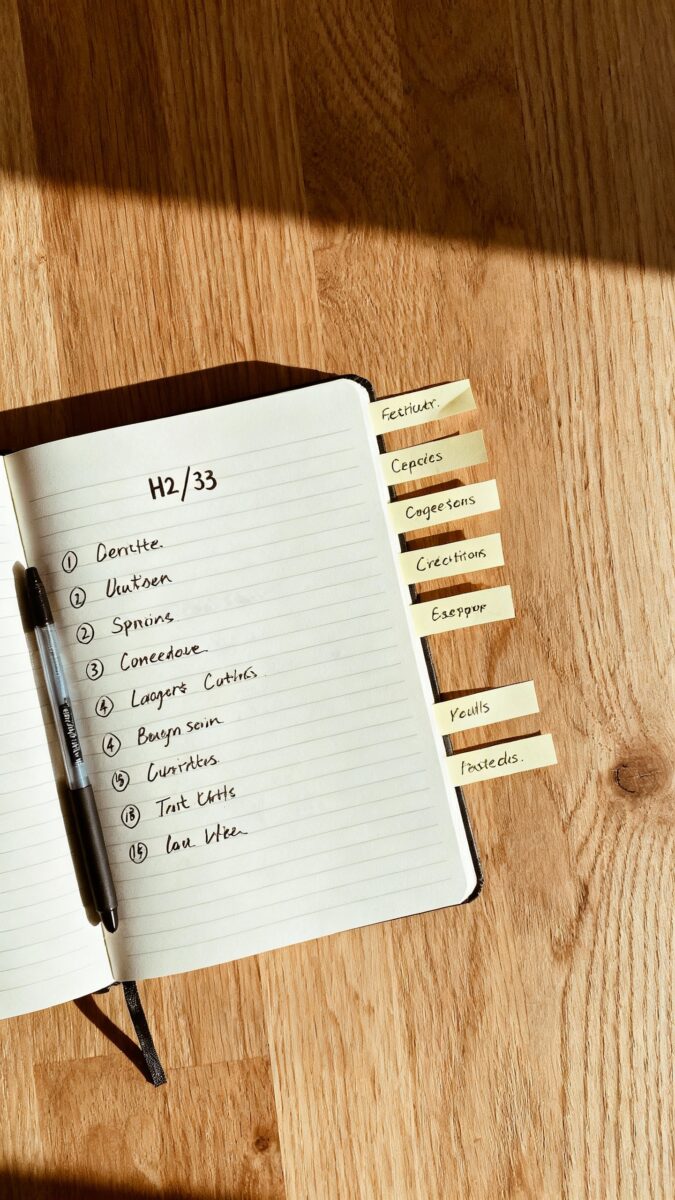 Overhead shot of notebook outline with H2/H3 bullets, black gel pen, sticky notes labeled subtopics,